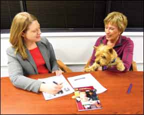 A woman sits at a conference table in an attorney's office; her dog is in her lap. She is consulting with an attorney, who is also seated at the table.