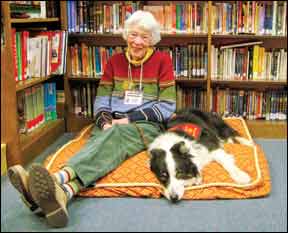 An older woman with white hair sits on a cushion at a library; her Border Collie is laying next to her. She wears an identification badge around her neck; she is a library volunteer in a "read to a dog" children's literacy program.