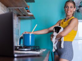 Woman, with a small dog in her arms cooking at stove while watching recipe on laptop.