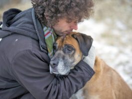Young Caucasian man wearing a winter coat and a scarf hugging his pet dog lovingly to comfort him as they are out for a walk through a snowy forest during winter. The dog is a mixed breed and brown and black in color with a sad expression on his face.