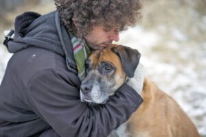 Young Caucasian man wearing a winter coat and a scarf hugging his pet dog lovingly to comfort him as they are out for a walk through a snowy forest during winter. The dog is a mixed breed and brown and black in color with a sad expression on his face.
