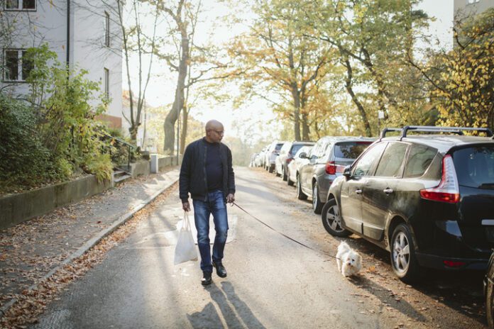 A fit older man walks a small dog white dog down neighborhood streets.